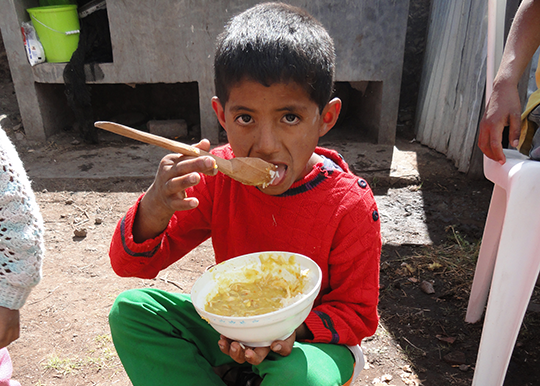 Peruvian boy sitting in dirt eating food out of a bowl with a wooden spoon